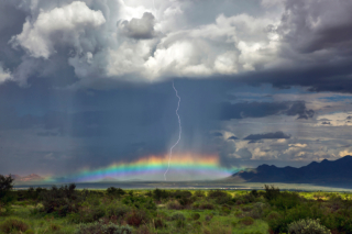 A storm over the high plains of New Mexico shadows a mountain range. A lightning bolt strikes through a rainbow.